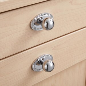 May include: Two polished chrome cabinet knobs featuring a spherical handle attached to an oval backplate. The backplate has a textured, diamond-patterned border. The knobs are mounted on light-colored wooden drawers with a visible grain pattern.