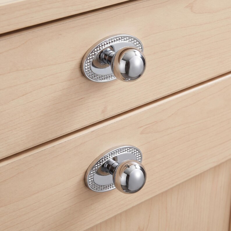 May include: Two polished chrome cabinet knobs featuring a spherical handle attached to an oval backplate. The backplate has a textured, diamond-patterned border. The knobs are mounted on light-colored wooden drawers with a visible grain pattern.