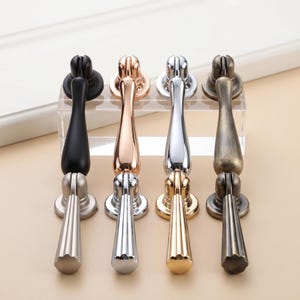 Peut inclure: Quatre ensembles de poignées de placard en métal disposés en rangée. Chaque ensemble comprend une poignée tombante verticale avec un sommet arrondi et un bouton plus petit assorti en dessous. Les finitions incluent noir mat, or rose, chrome poli et laiton antique.