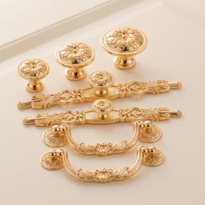 May include: A collection of gold-toned metal cabinet hardware including three round knobs of varying sizes, two elongated drawer pulls with ornate filigree detailing, and two curved drop-style drawer handles with matching circular mounting plates.