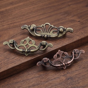 Pode incluir: Três puxadores de gaveta de metal com placas traseiras ornamentadas e vazadas e alças oscilantes. Dois puxadores apresentam um acabamento em latão antigo, enquanto o terceiro tem um acabamento em tom de cobre. Cada peça exibe volutas complexas.