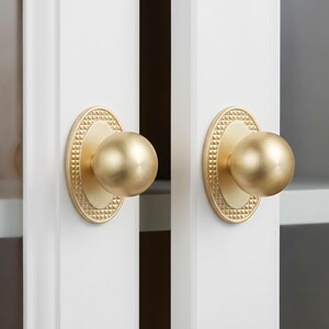 May include: Two matching brass door knobs with a spherical handle and an oval backplate. The backplate features a textured, pyramid-like pattern around the perimeter, while the central knob has a smooth, brushed gold-tone finish.