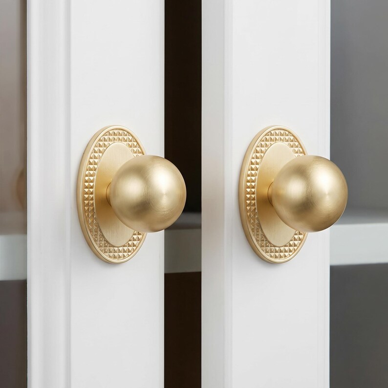 May include: Two matching brass door knobs with a spherical handle and an oval backplate. The backplate features a textured, pyramid-like pattern around the perimeter, while the central knob has a smooth, brushed gold-tone finish.