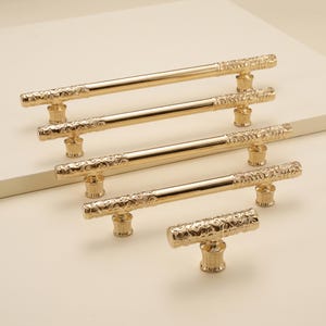 Op de afbeelding: Een collectie van vijf goudkleurige kastgrepen en één T-vormige knop. Elk stuk heeft een gepolijst middengedeelte en getextureerde, sierlijke uiteinden met ingewikkelde wervelpatronen. Het beslag is in een verspringende formatie geplaatst.
