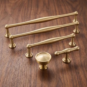 May include: A collection of five brass cabinet hardware pieces featuring a brushed gold finish. The set includes three elongated bar pulls with a central tapered design, one circular knob with a flat top, and one small T-shaped handle.