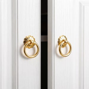 Op de afbeelding: Twee goudkleurige metalen ringgrepen bevestigd op witte kastdeuren. Elke greep heeft een cirkelvormige ring die hangt aan een decoratieve, geschulpte, bloemvormige achterplaat. De kastdeuren hebben een verticale houtnerf en een paneelontwerp.