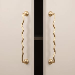 Puede incluir: Dos tiradores de armario verticales con una silueta ondulada y serpenteante. Estas piezas de herraje presentan un acabado dorado mate y liso, montadas en los bordes de dos puertas de armario claras, cada una asegurada por placas base circulares.