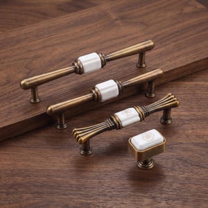 May include: Three brass-toned metal cabinet pulls and one matching cabinet knob. Each piece features a central white ceramic block. The bottom pull and knob display a delicate, thin-lined floral pattern etched onto the white ceramic surface.