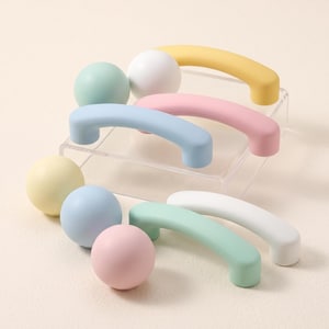 Pode incluir: Uma coleção de ferragens para móveis, incluindo puxadores esféricos e alças de gaveta arqueadas em cores pastel. O conjunto apresenta acabamentos foscos em tons de verde menta, branco, amarelo, azul claro e rosa suave sobre um suporte.