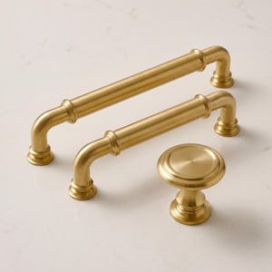 May include: Two brushed brass cabinet pulls with a cylindrical bar design and a matching round cabinet knob. Each piece features a smooth, metallic finish with a warm gold tone, rounded ends, and a sturdy base for mounting to furniture or cabinetry.