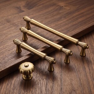 May include: A collection of brass-toned cabinet hardware including three elongated bar pulls with fluted end caps and one matching round knob. Each piece features a smooth, matte metallic finish and a consistent design with ribbed detailing.