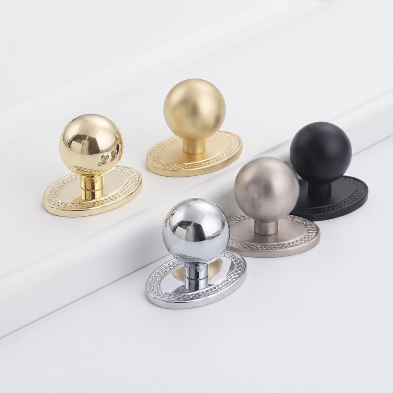 May include: Five cabinet knobs with spherical heads and oval base plates featuring a beaded edge. The set includes finishes in polished gold, matte gold, polished chrome, brushed nickel, and matte black, each mounted on a short cylindrical stem.