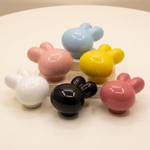 May include: A collection of six ceramic cabinet knobs shaped like stylised rabbit heads. The knobs feature a smooth, glossy finish and come in a variety of solid colours including white, light blue, yellow, black, and two shades of pink.