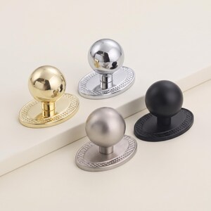 May include: Four cabinet knobs with spherical heads and oval base plates featuring a textured border. The set includes one polished gold, one polished chrome, one matte silver, and one matte black finish, each with a central stem connecting the knob.