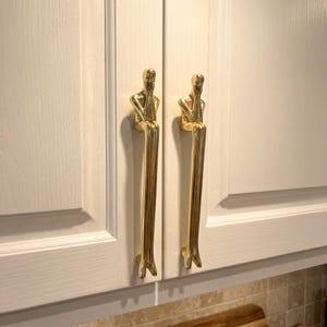 May include: Two gold-colored metal cabinet pulls shaped like elongated human figures in a seated position. Each handle features a smooth, reflective finish and is mounted vertically on a pair of off-white cabinet doors with a wood grain texture.
