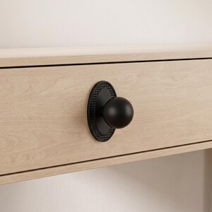 May include: A matte black spherical cabinet knob mounted on an oval backplate with a textured, pyramid-patterned border. The hardware is attached to a light-toned, wood-grain drawer front, showcasing a clean, functional design for furniture.