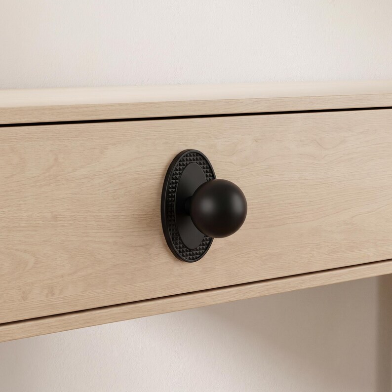 May include: A matte black spherical cabinet knob mounted on an oval backplate with a textured, pyramid-patterned border. The hardware is attached to a light-toned, wood-grain drawer front, showcasing a clean, functional design for furniture.