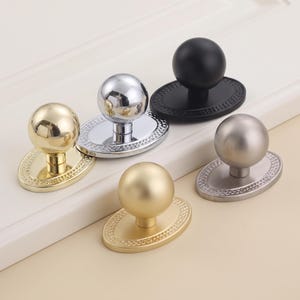 May include: Five spherical cabinet knobs with oval base plates featuring a textured border. The set includes one polished gold, one polished chrome, one matte black, one brushed gold, and one brushed nickel finish, all arranged on a white surface.