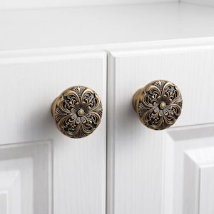 May include: Two round cabinet knobs with an antique brass finish, featuring a raised, intricate floral scrollwork pattern with dark recessed detailing. The knobs are mounted on white cabinet doors with a visible wood grain texture.