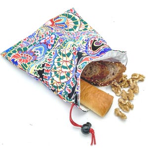 Pode incluir: Um saco de pão reutilizável em tecido estampado com fecho de cordão. O saco contém um pão, um pãozinho e nozes. O design do saco apresenta um padrão floral e paisley colorido com detalhes em vermelho, azul e verde.