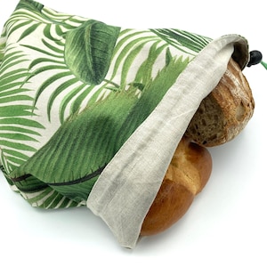 Op de afbeelding: Een stoffen tas met trekkoord voorzien van een groen en wit tropisch palmbladpatroon, gevuld met twee broden. De tas heeft een natuurlijke beige linnenkleurige voering en een zwarte plastic koordstopper aan de zijkant.