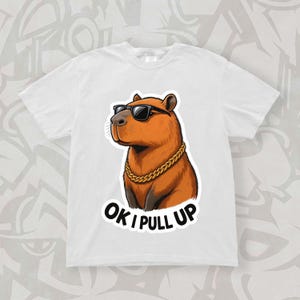 May include: White t-shirt featuring a cartoon capybara wearing sunglasses and a gold chain. The capybara has brown fur and is set against a white background. The text "OK I PULL UP" is printed below the capybara.