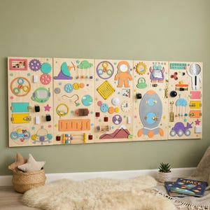 Pu&ograve; includere: Una grande lavagna in legno con vari elementi interattivi per bambini. Include ingranaggi, chiusure, un labirinto, un orologio e un razzo. La lavagna &egrave; montata su una parete verde chiaro, con un cesto di stelle e un tappeto in primo piano.