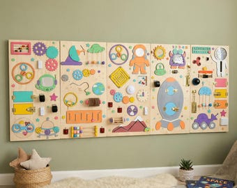 May include: A large wooden busy board with various interactive elements for children. Features include gears, latches, a maze, a clock, and a rocket ship. The board is mounted on a light green wall, with a basket of stars and a rug in the foreground.