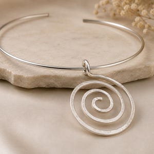 Puede incluir: Un collar tipo gargantilla de metal plateado con un gran colgante en forma de espiral circular. El colgante cuelga de un fino aro de alambre abierto. La superficie del colgante en espiral tiene un acabado martillado y texturizado.