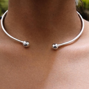 Könnte beinhalten: Eine silberfarbene Choker-Halskette mit schlichtem, modernem Design. Die Halskette hat einen gebogenen Stab, der um den Hals liegt, mit einer kleinen, runden Kugel an jedem Ende. Der Schmuck wird vor einem hautfarbenen Hintergrund gezeigt.