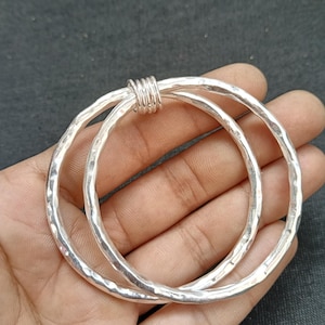 May include: Two silver bangles with a hammered texture are connected by a wrapped silver wire. The bangles are round and have a slightly irregular shape. The jewelry is displayed on a hand against a dark background.