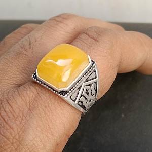 Puede incluir: Un anillo de plata con una gran piedra preciosa amarilla cuadrada. El anillo tiene un diseño ornamentado en la banda y un engaste de bisel plateado alrededor de la piedra. La piedra preciosa es de color amarillo brillante y soleado.