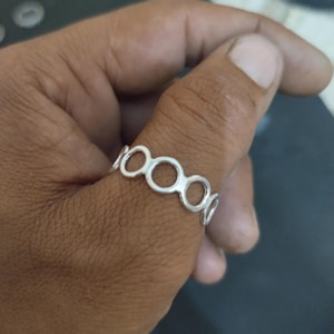 May include: A silver ring featuring a series of connected open circles. The ring is worn on a finger, showcasing its delicate design and polished finish. The ring's circular pattern creates a modern and minimalist aesthetic.