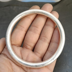 May include: A silver-colored bangle bracelet with a smooth, rounded design. The circular bracelet is held in a hand, showcasing its polished surface and simple, elegant style. The bangle is a classic jewelry piece.