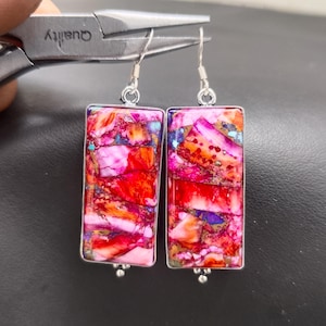 May include: A pair of rectangular earrings with a vibrant, multicolored stone inlay. The earrings feature a silver-colored metal frame and ear hooks. The stone's colors include shades of red, pink, orange, and blue. The earrings are approximately 1.5 inches long.