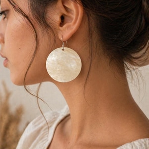May include: Cream-coloured, circular earrings with a pearlescent finish. The earrings are suspended from small silver hooks. The image shows a close-up of the earrings against a person's skin and clothing.