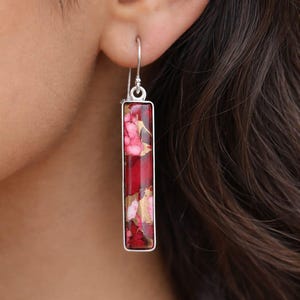 May include: A rectangular silver earring with a red, pink, and gold stone. The stone has a marbled appearance. The earring is hanging from a silver hook. The earring is worn on a person's ear.
