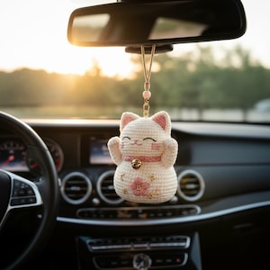 Może przedstawiać: Kremowa, szydełkowana ozdoba w kształcie kota zawieszona na lusterku wstecznym. Kot ma różowe wnętrza uszu, różową obrożę z małym złotym dzwonkiem oraz różowy haft kwiatowy na brzuchu z akcentami w kształcie złotych gwiazdek.