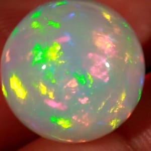 May include: A spherical opal gemstone with a milky white base. The opal exhibits flashes of green, yellow, and pink. The polished surface reflects light, creating a vibrant display of colour. The gemstone is a beautiful example of natural iridescence.
