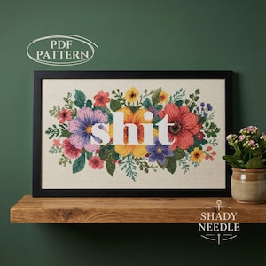 Może przedstawiać: Wzór kwiatowy haftu krzyżykowego ze słowem "shit" w białych literach blokowych. Kwiaty są w odcieniach różu, fioletu, żółci i czerwieni. Projekt jest oprawiony w czarną ramę.
