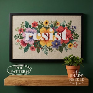 Può includere: Opera d'arte a punto croce incorniciata con la parola "resist" in lettere bianche, circondata da una vivace composizione floreale nei toni del rosa, giallo, rosso e viola. L'opera è esposta su una parete verde sopra una mensola di legno.