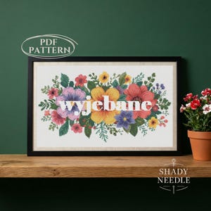 Op de afbeelding: Een ingelijst kruissteek bloemenontwerp met het woord "Wyjebane" in het wit. De bloemen zijn in tinten roze, geel, paars en rood. De lijst is zwart en de achtergrond is donkergroen.