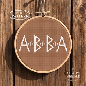 May include: A circular wooden embroidery hoop containing brown fabric with white cross-stitched letters spelling A+B+B+A. The text PDF PATTERN is visible in the upper left corner, and the text SHADY NEEDLE is visible in the lower right corner.