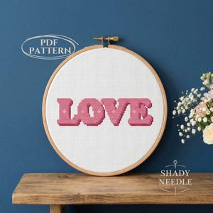 May include: A cross-stitch embroidery hoop with the word "LOVE" in pink block letters. The design is on white fabric, set in a wooden hoop. The text "PDF PATTERN" is in a white oval at the top left. The brand name "SHADY NEEDLE" is at the bottom right.