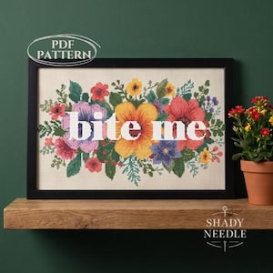 May include: A framed cross-stitch embroidery featuring a vibrant floral arrangement of red, yellow, purple, and green flowers surrounding the white text "bite me" centred on a light fabric background, with a PDF PATTERN label at the top.