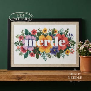 Könnte beinhalten: Schwarz gerahmtes Kreuzstichmuster mit dem Wort "merde" in weißen Buchstaben, umgeben von einem lebendigen Blumenarrangement. Die Blumen sind rot, gelb, lila und rosa mit grünem Laub. Die Worte "PDF PATTERN" befinden sich oben links.