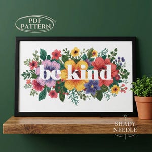 May include: A framed cross-stitch floral design with the words "be kind" in white. The flowers are in shades of pink, purple, yellow, and red, with green leaves. The frame is black, and the image is on a wooden shelf.