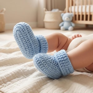 May include: A pair of light blue baby booties crafted from thick yarn with a visible crochet stitch pattern. The booties feature a folded cuff design and a rounded toe shape, providing a textured appearance suitable for infant footwear.