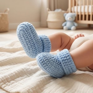 May include: A pair of pale blue crochet baby boots. The boots are made of soft yarn and have a rolled cuff. The baby is lying on a cream-coloured blanket. The background includes a cot, a teddy bear, and a window.