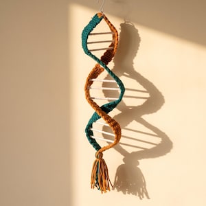 May include: A hanging crochet DNA double helix model featuring two intertwined strands in teal and mustard yellow yarn. The structure is connected by horizontal white bars and terminates in a decorative tassel at the bottom, suspended by a metal hook.